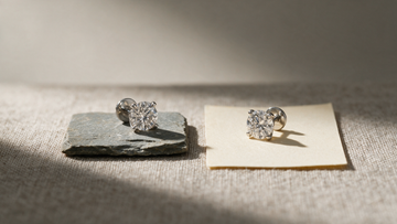 Two identical diamond solitaire studs placed side by side for comparison, one on slate and one on paper