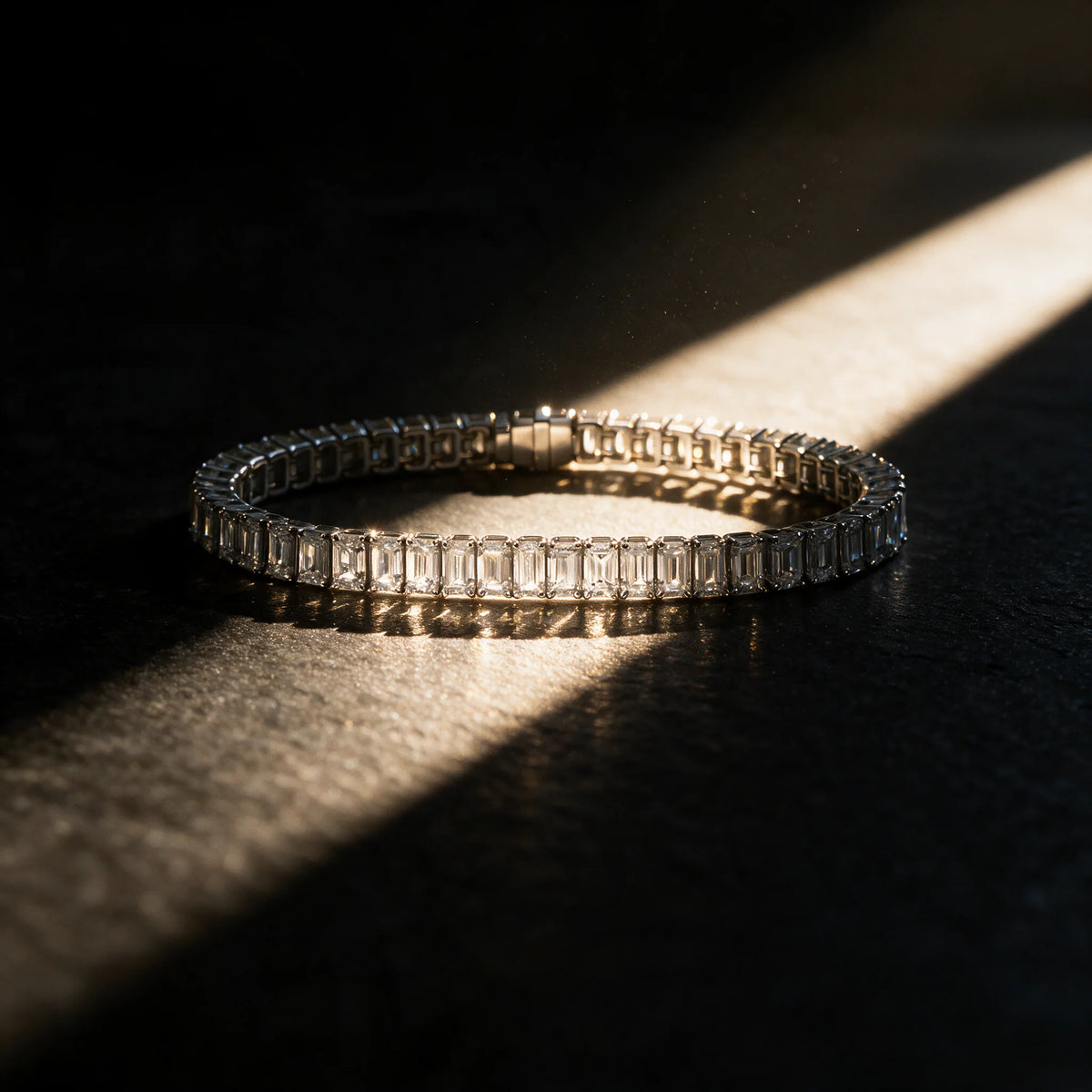 Carat Club Co. Meridian men's emerald-cut tennis bracelet laid in a circle on dark slate, a diagonal shaft of warm light crossing the white gold box clasp and the underside gallery.