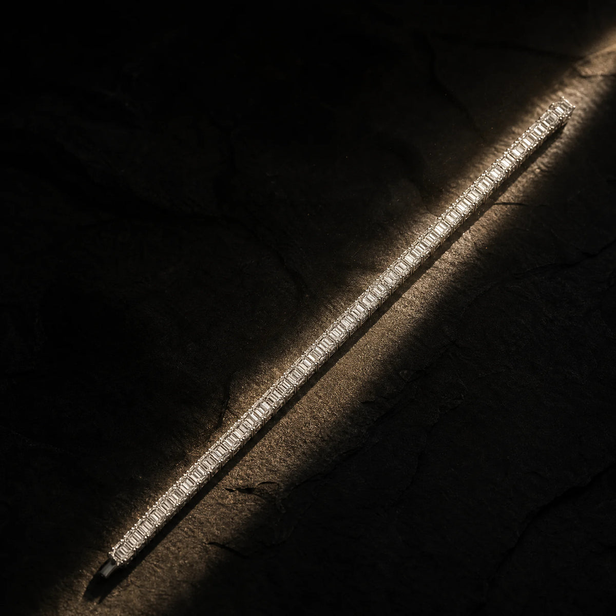 Meridian men's emerald-cut tennis bracelet laid full length in a shaft of light on slate, Carat Club Co.