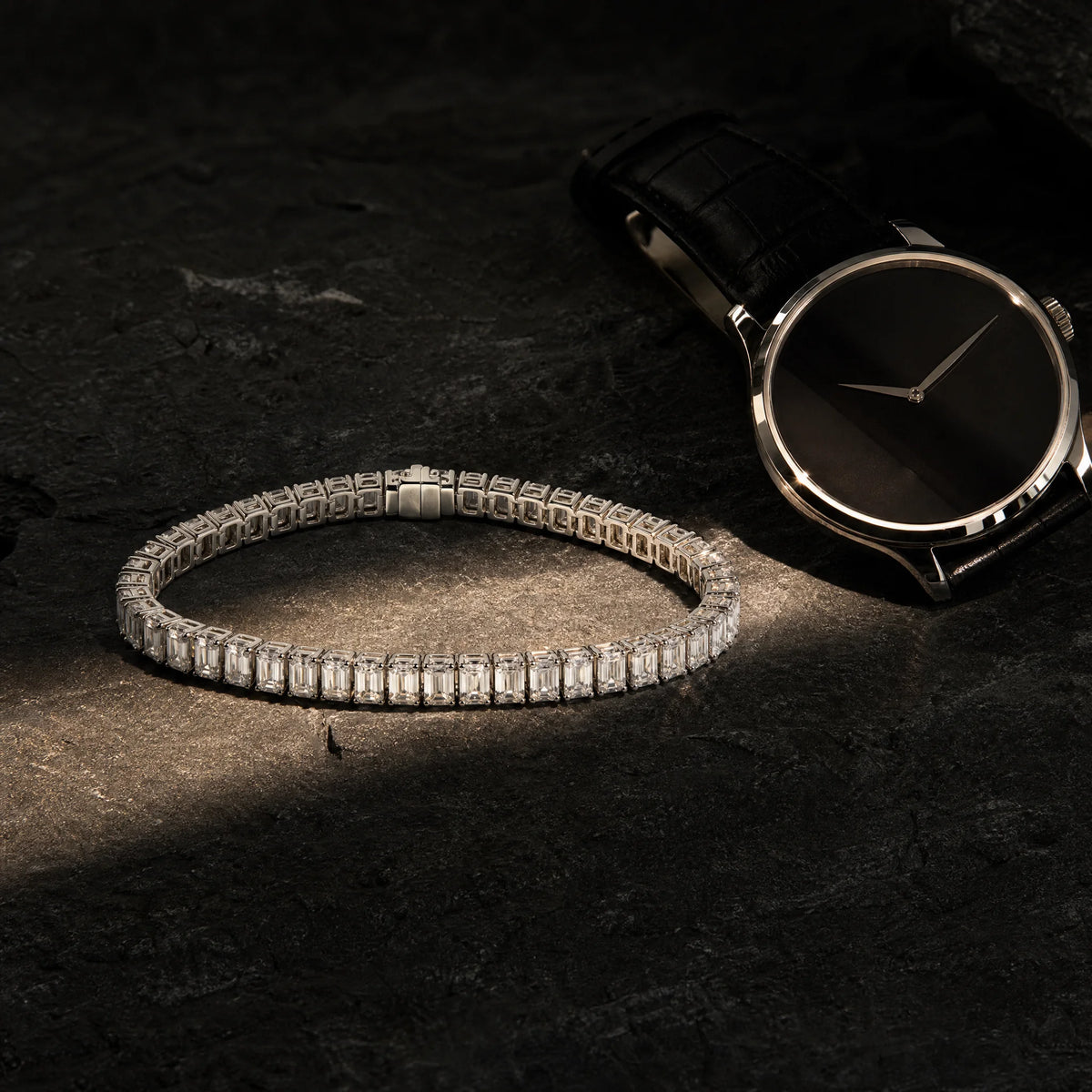 Carat Club Co. Meridian men's emerald-cut tennis bracelet in white gold resting on dark slate beside a black-strap dress watch, warm spotlight men's styling shot.